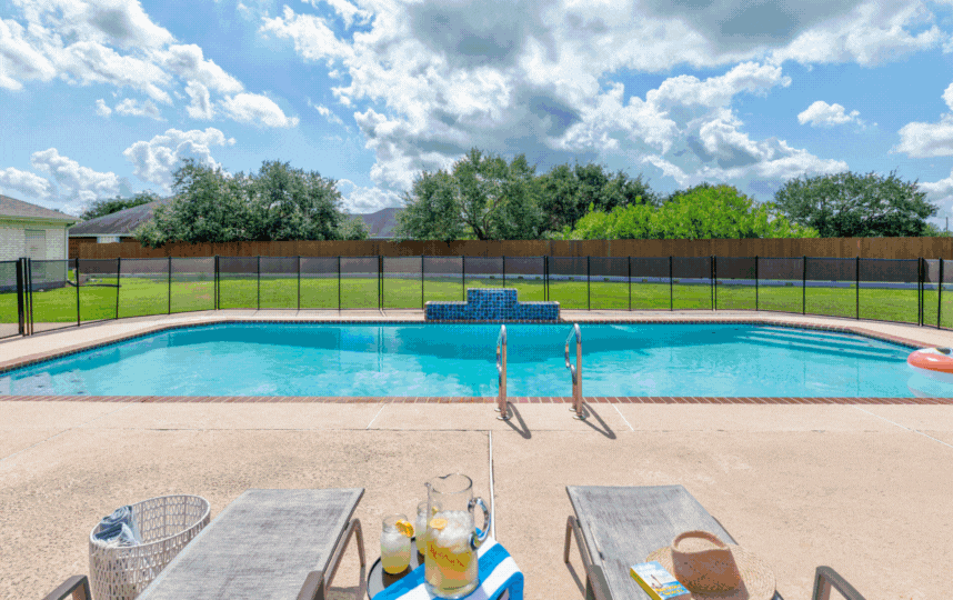 Outdoor swimming pool with a safety fence, lounge chairs, and a table with lemonade.