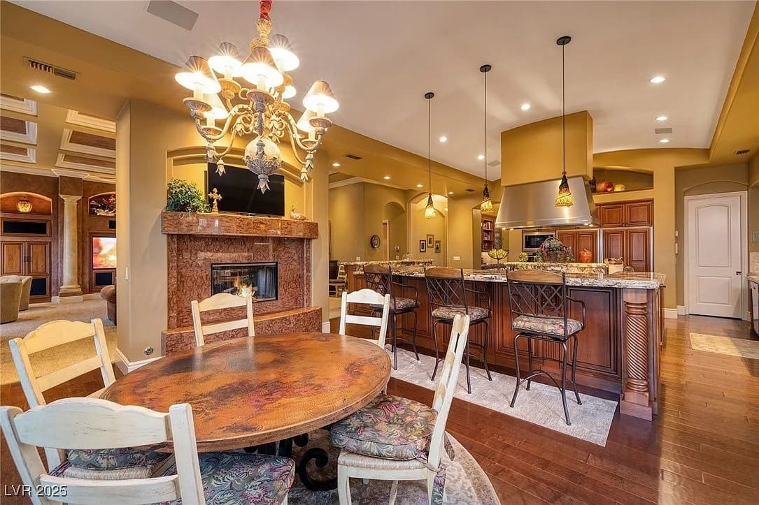 Elegant kitchen and dining area with wooden furniture, chandelier, and marble fireplace. Warm lighting.
