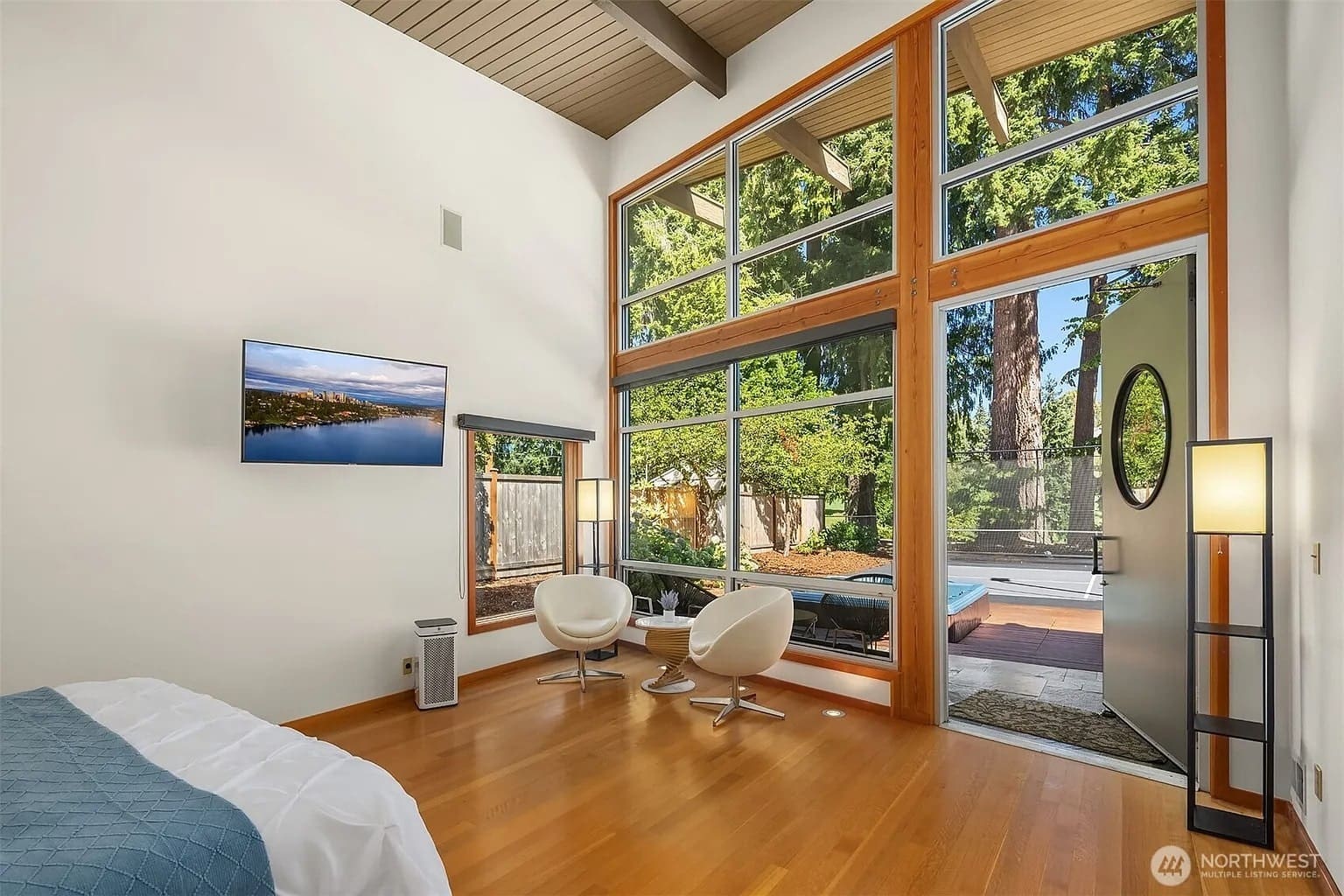 Modern bedroom with large windows, two chairs, wall-mounted TV, and view of outdoor patio and trees.