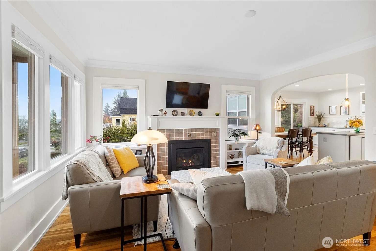 Bright living room with beige sofas, fireplace, wall-mounted TV, and view of dining area and kitchen.