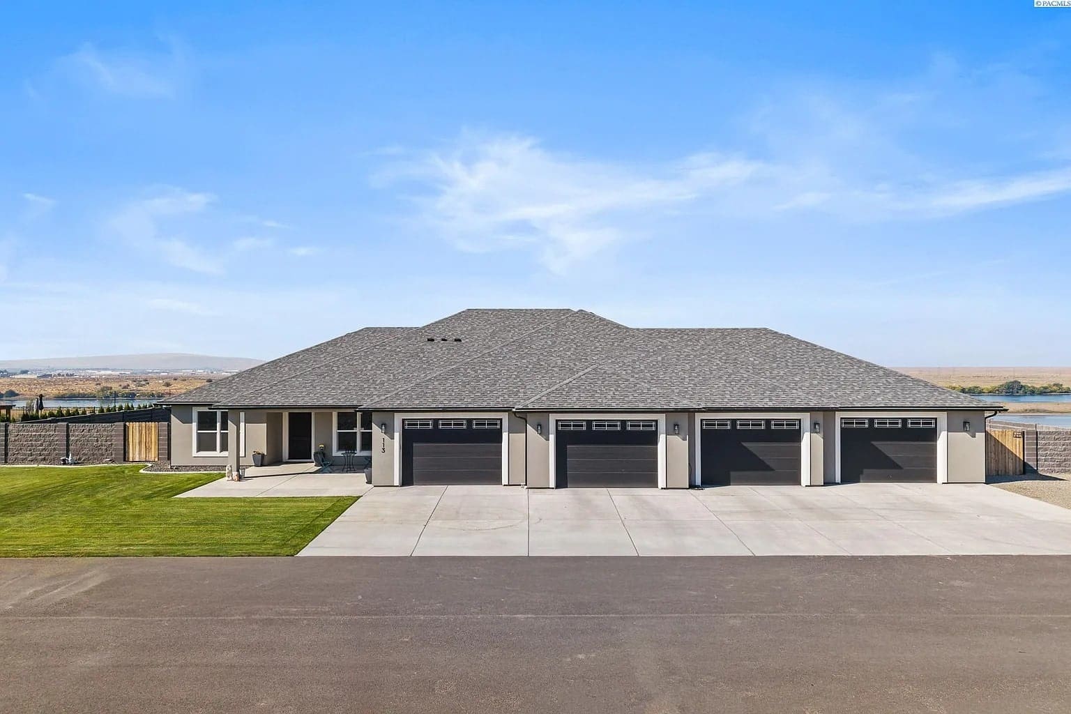 Large modern house with a four-car garage, gray roof, and surrounding lawn under a clear blue sky.
