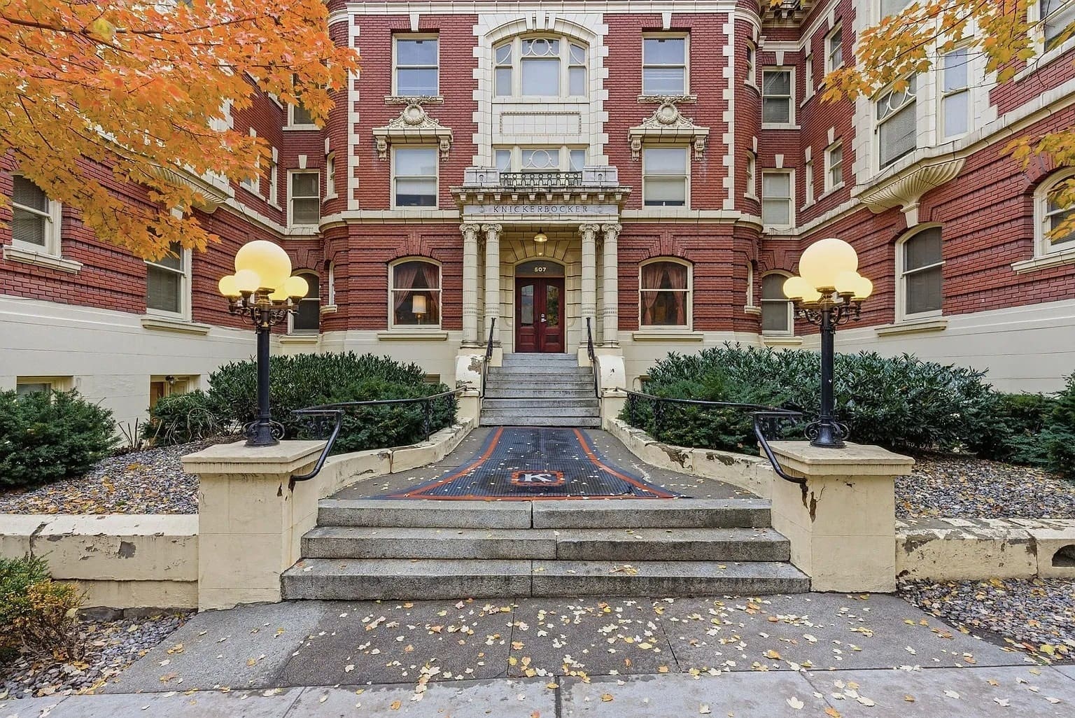 Historic red brick building entrance with ornate details, surrounded by autumn trees and lamps.
