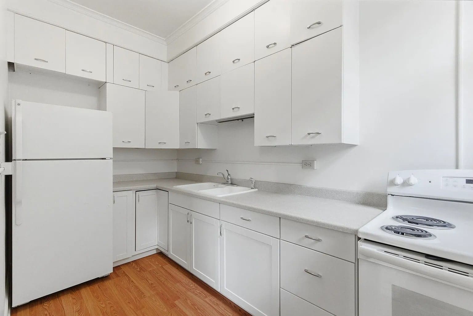 White kitchen with wooden floor, featuring cabinets, a refrigerator, sink, and electric stove.