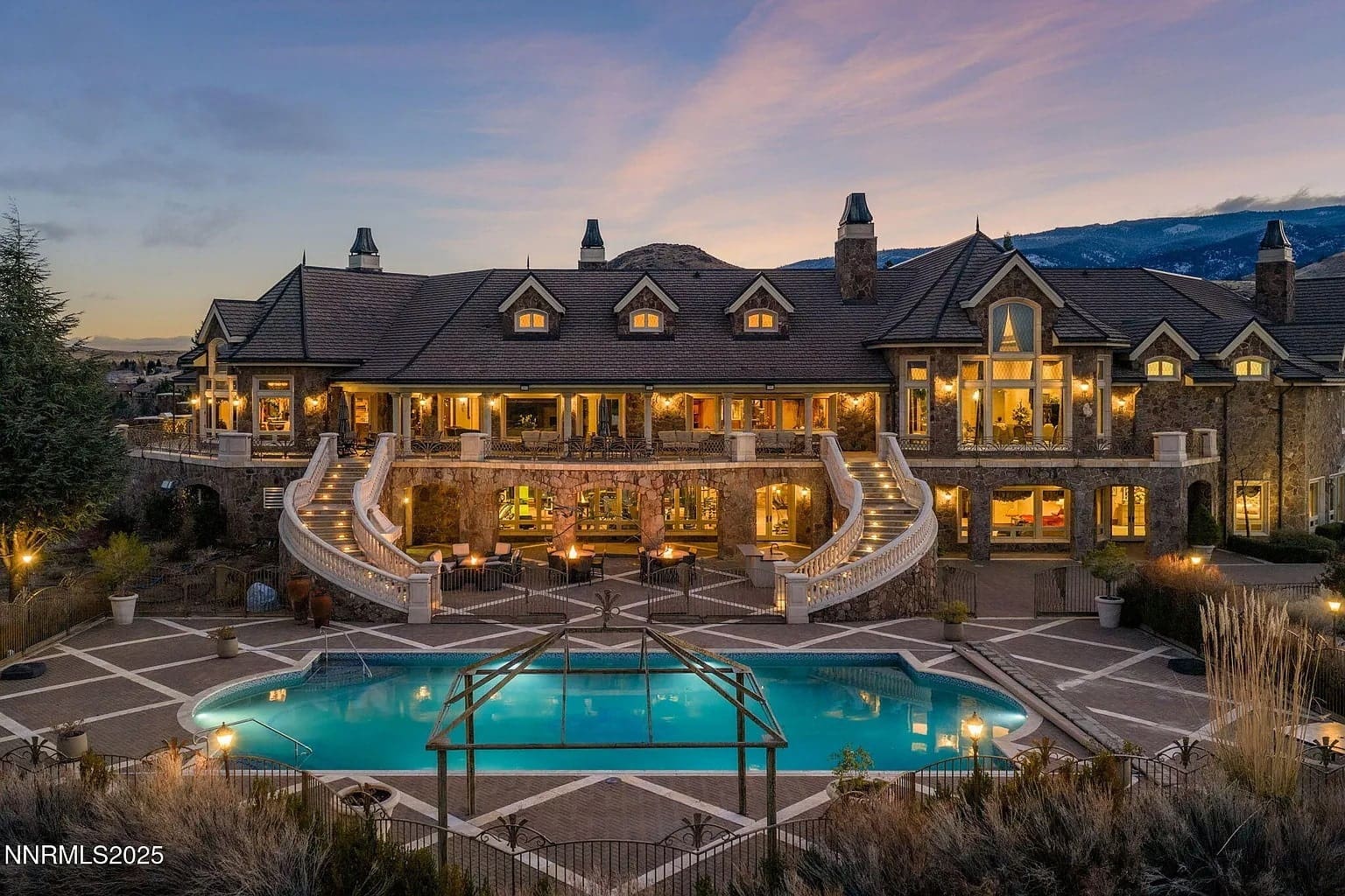 Large stone mansion with multiple staircases, lit windows, and a pool at dusk.