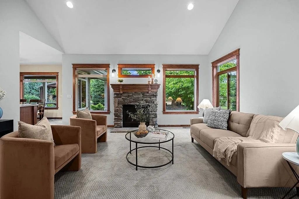 Living room with beige sofa, two brown chairs, stone fireplace, and large windows overlooking greenery.
