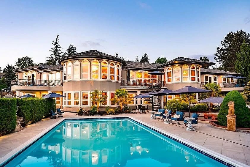 Luxurious mansion with large windows, surrounded by trees, featuring a spacious pool and patio area.