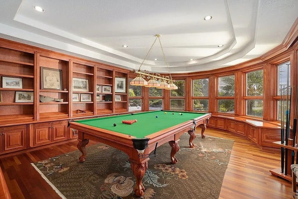 Luxurious room with a billiards table, wooden shelves, and large windows overlooking greenery.