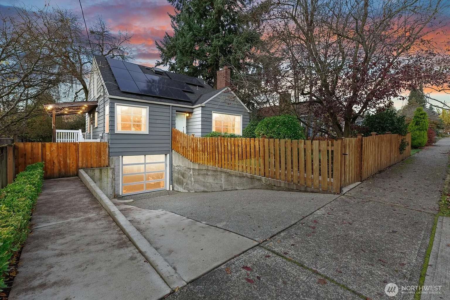 Small gray house with solar panels, driveway, and wooden fence at sunset.