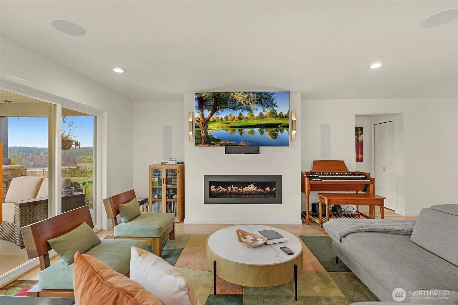 Modern living room with a wall-mounted TV, fireplace, and large windows overlooking a scenic view.